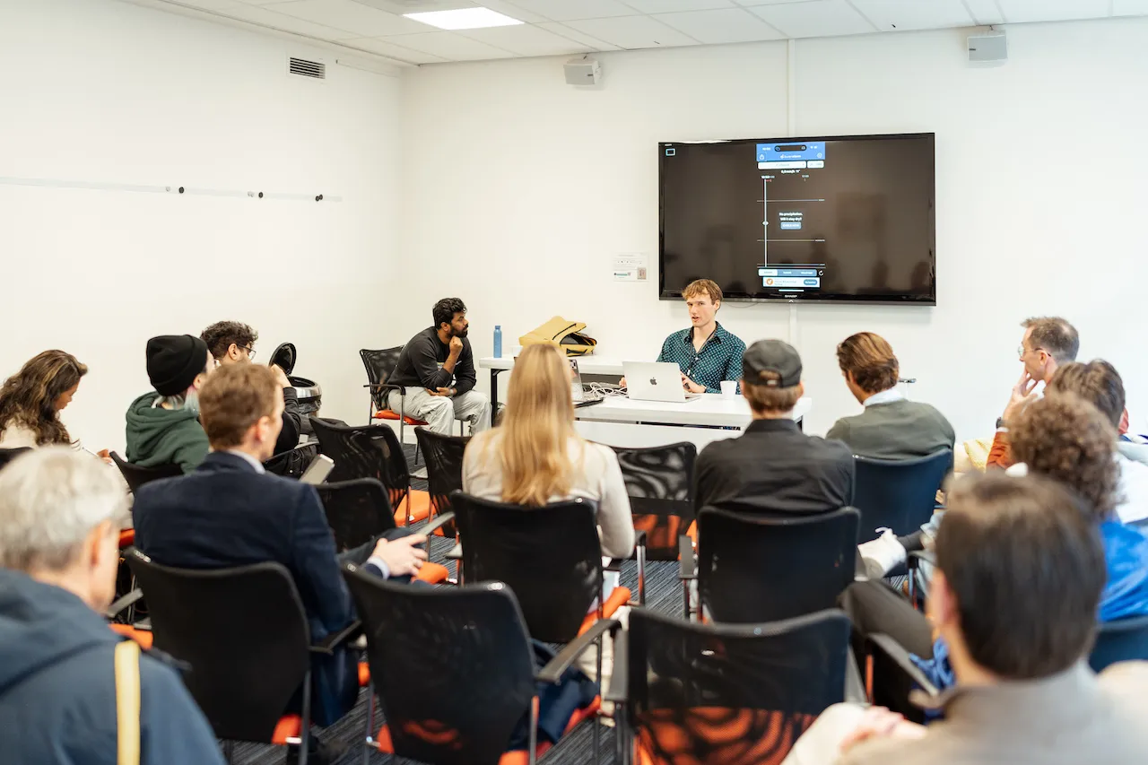 A group of attendees seated in a workshop setting, focusing on a presenter at the front of the room. The presenter, Jesse Wienholts, is seated at a table with a laptop, facing the audience. A large screen behind the presenter displays a flowchart or dialogue tree. The room appears well-lit with white walls and a casual, collaborative atmosphere.
